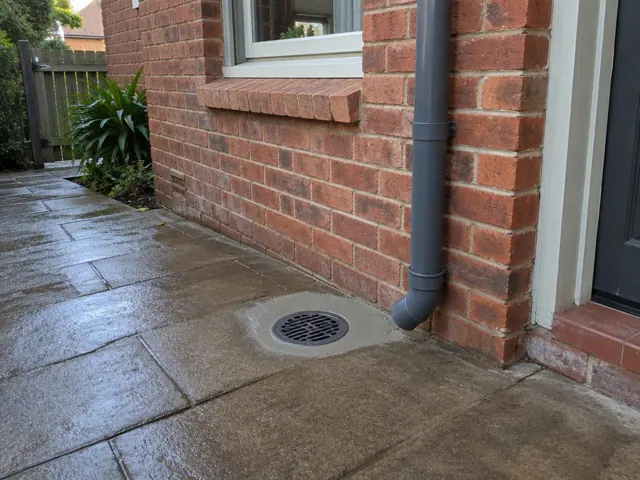 Outdoor drain grate beside brick house wall and wet paved patio in Abbotsford