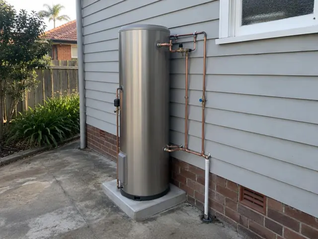Outdoor hot water cylinder beside weatherboard wall and brick base in a suburban yard in Haberfield